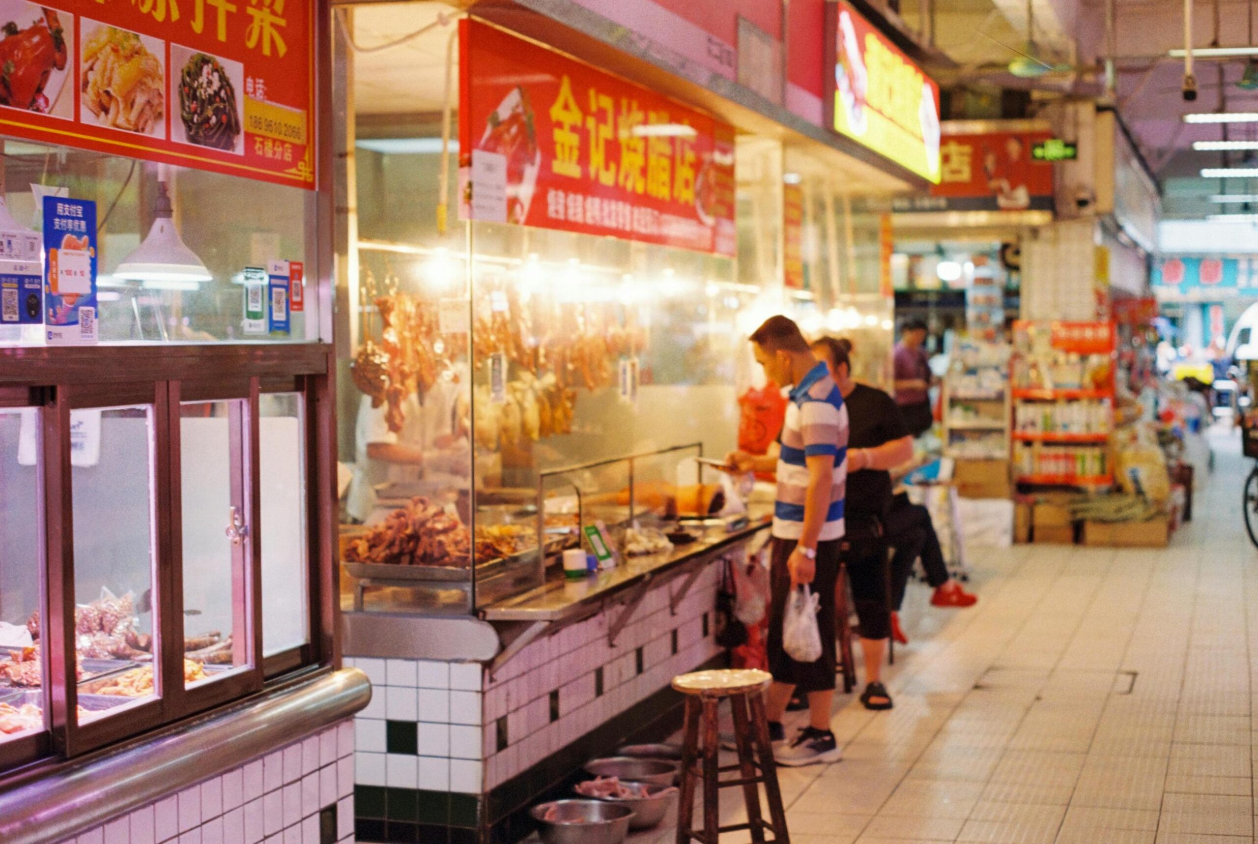 This vibrant indoor market scene features a food stall displaying hanging roasted meats behind a glass counter, illuminated by bright overhead lights. A customer in a striped shirt waits near the front, while the tiled aisle extends back past other shops and shelves of goods.