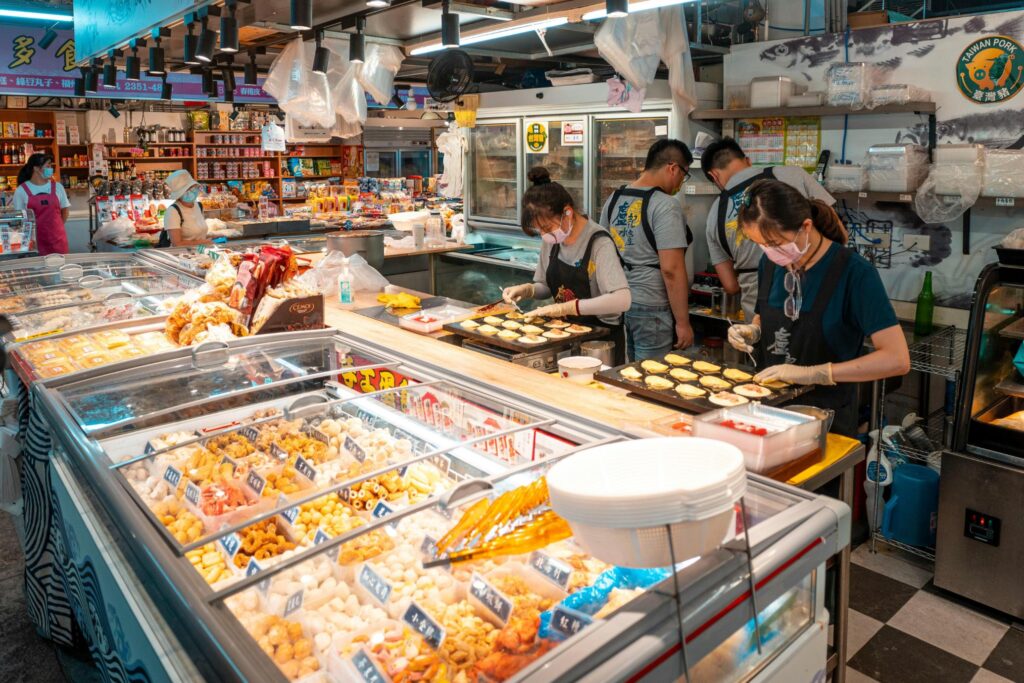 This image depicts a bustling market stall where several masked workers in aprons are actively cooking on griddles behind a counter. In the foreground, large glass display freezers are packed with a wide assortment of frozen ingredients like fish balls and dumplings, while shelves of packaged groceries line the background.
