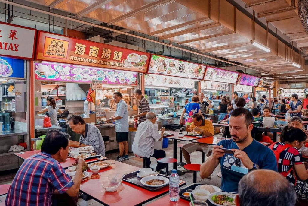 This busy hawker center scene features a row of food stalls with bright signage, including a prominent Hainanese Chicken Rice vendor, serving a crowded dining area. Patrons are seated at communal red tables enjoying their meals, while a man in the foreground captures a photo of his dish with a smartphone.