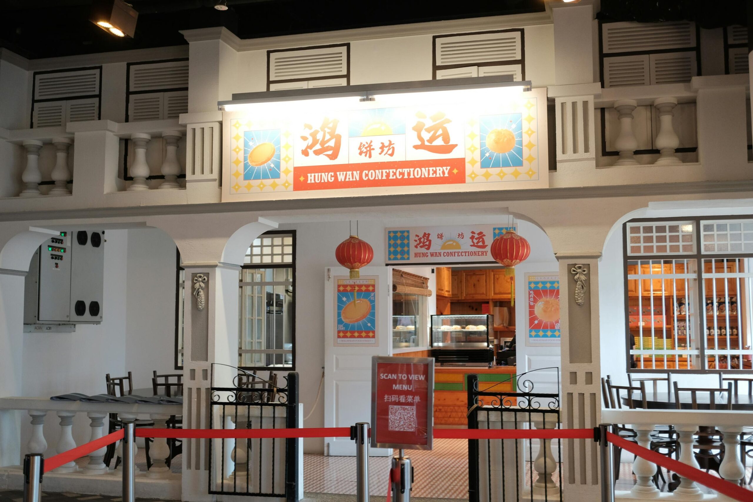 This image displays the facade of "Hung Wan Confectionery," designed with white architectural details like columns and balustrades reminiscent of a traditional shophouse. Red lanterns hang above the entrance behind a red rope barrier, where a standing sign instructs visitors to scan a code to view the menu.