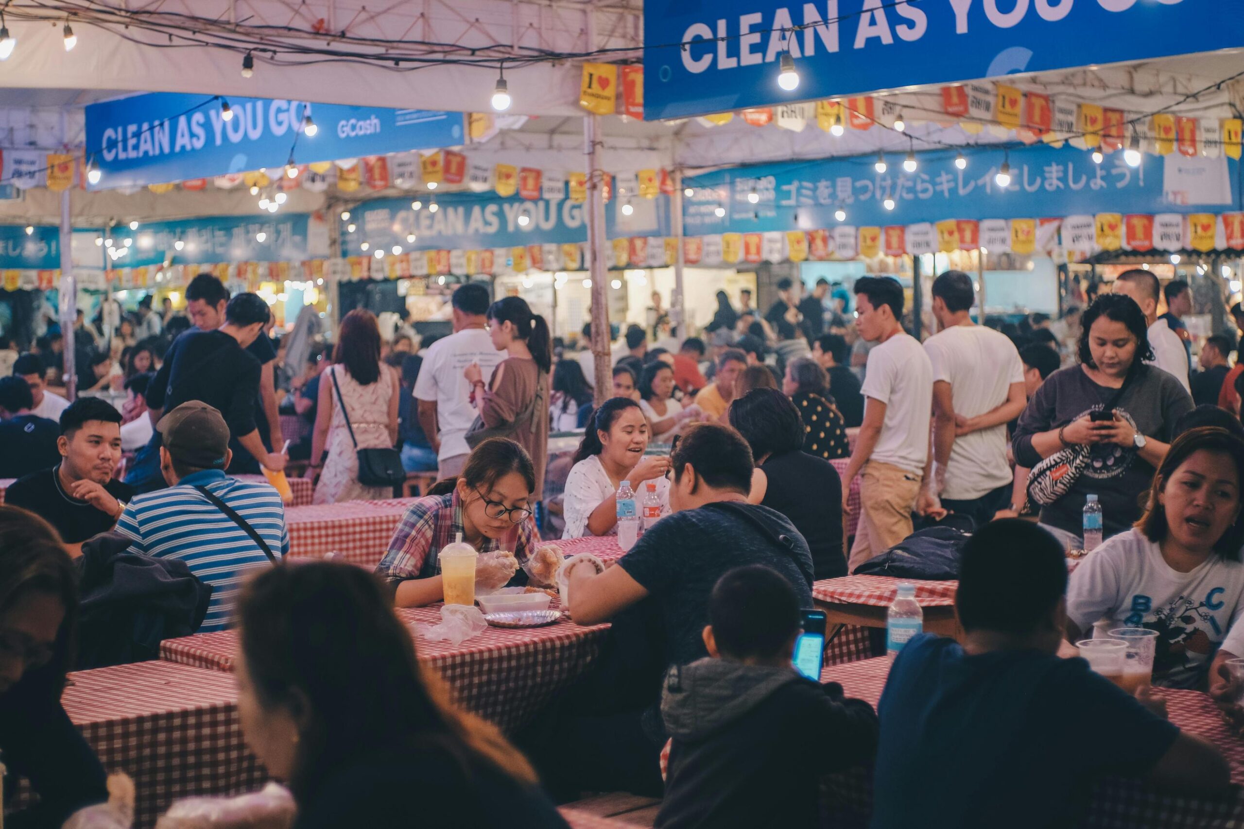 This image captures a bustling night market scene where a large crowd is seated at long tables covered in red and white checkered tablecloths. String lights hang from the tent ceiling above the diners, illuminating blue banners that prominently display the slogan "CLEAN AS YOU GO" alongside other branding.