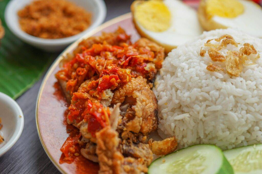 A high-angle close-up of a plate featuring a mound of white rice topped with fried shallots. It is served with crispy fried chicken smothered in a bright red sambal chili sauce, fresh cucumber slices, and a halved hard-boiled egg, with small side dishes of condiments visible in the background.