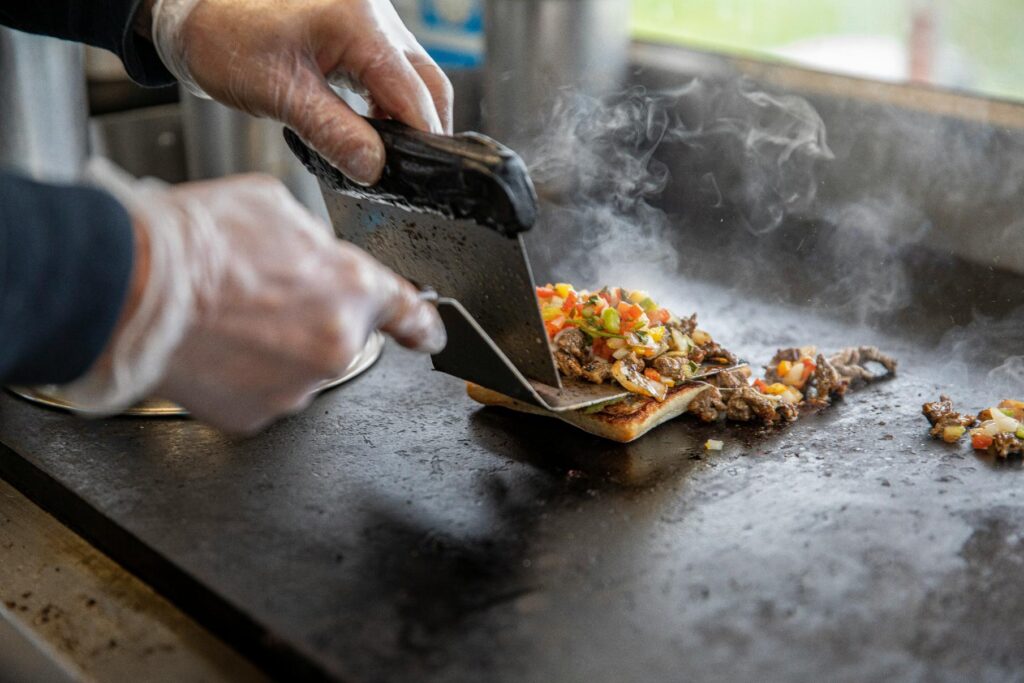 A pair of gloved hands uses a metal scraper to lift a steaming portion of chopped meat and peppers onto a slice of toasted bread on a flat-top grill. The close-up captures the sizzling action as smoke rises from the savory mixture, highlighting the fresh preparation of the meal.