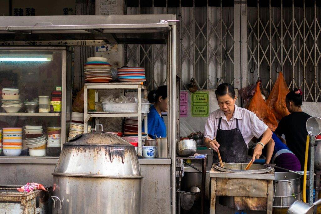 An outdoor hawker food stall where a woman in an apron is cooking at a large steaming pot with long wooden chopsticks. The stall is filled with stacks of colorful bowls and plates, large metal steamers, and set against a backdrop of traditional accordion-style metal shop doors.