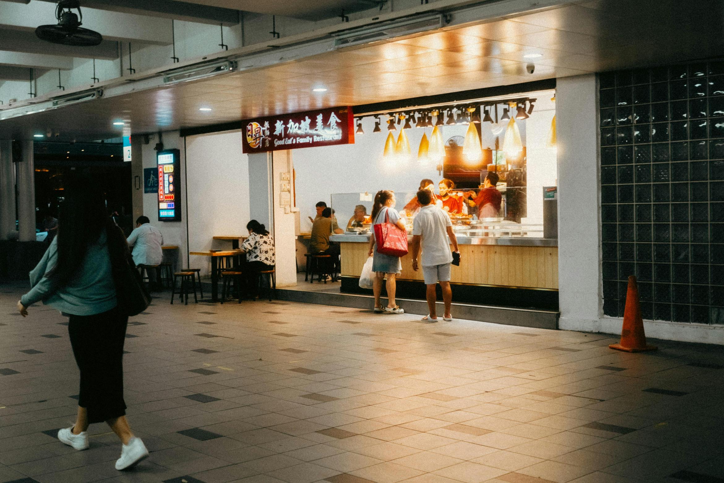 This image features a brightly illuminated food stall labeled "Good Eat's Family Restaurant," where customers are ordering at the counter and dining at nearby tables. A pedestrian walks across the tiled foreground in the dim open space, which is framed by a glass block wall and an orange safety cone on the right.