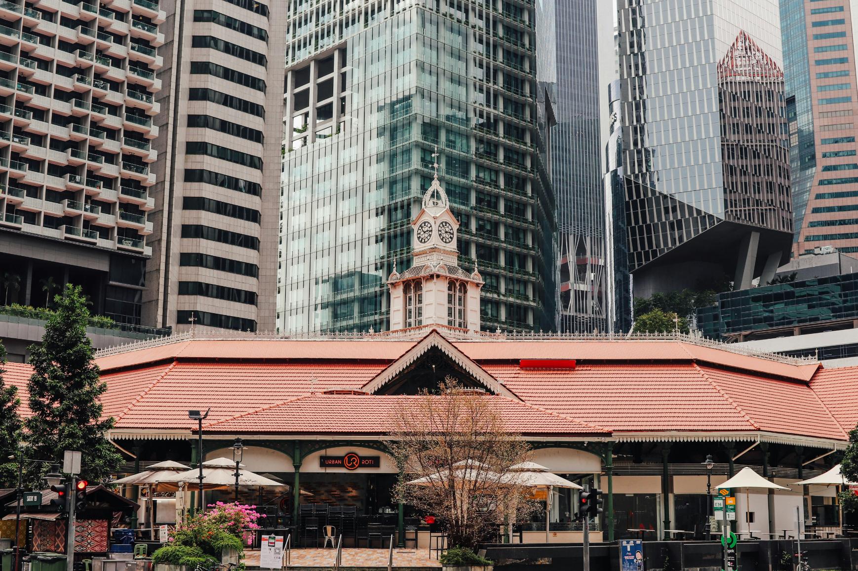 This image features the historic Lau Pa Sat market building, distinguished by its expansive red-tiled roof and central Victorian clock tower, standing in stark contrast to the towering modern skyscrapers that surround it. The low-rise structure is nestled within a dense urban landscape of glass and steel office buildings, showcasing a unique blend of heritage architecture and contemporary city development.