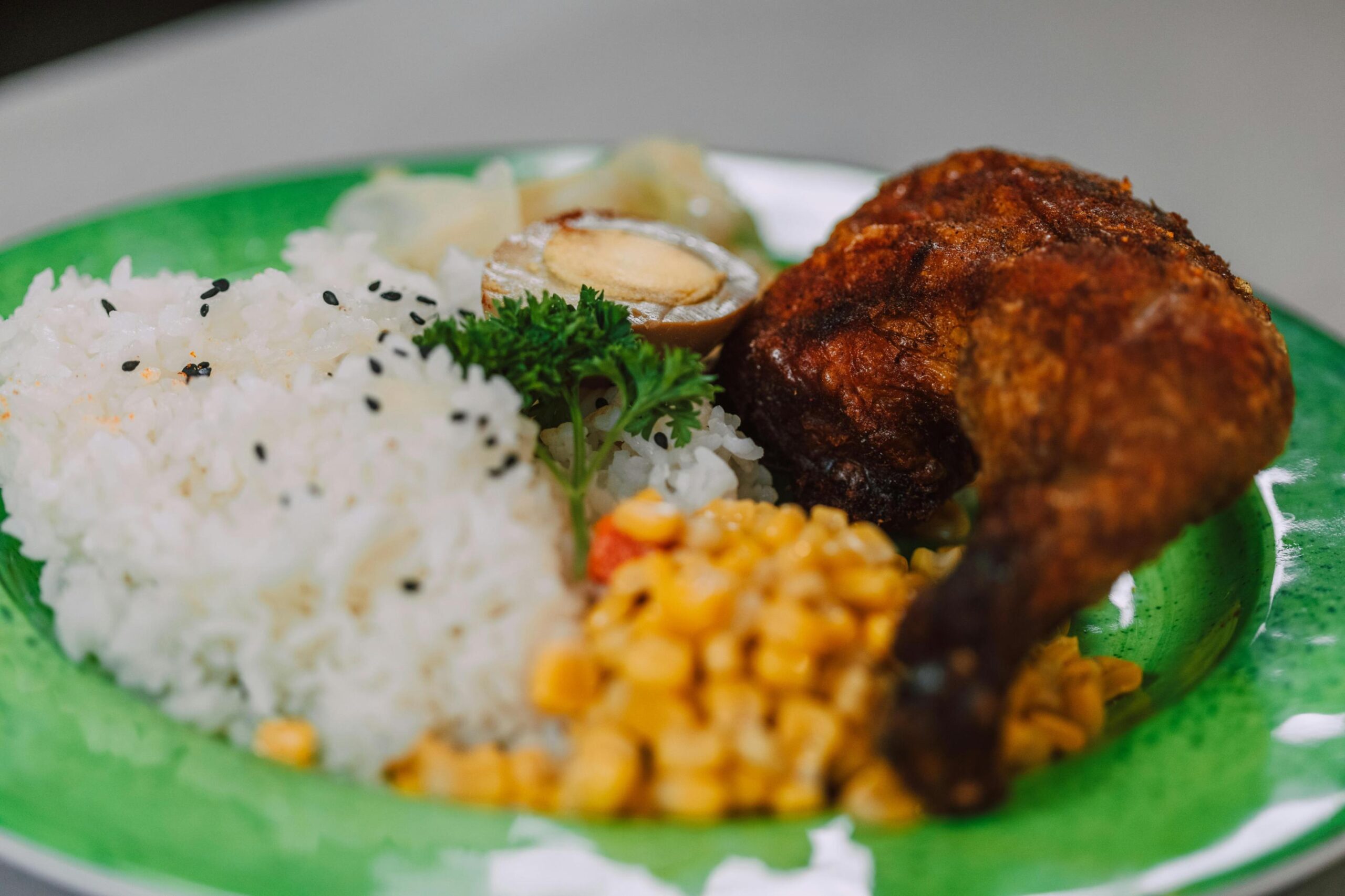 A bright green plate features a serving of white rice sprinkled with black sesame seeds, accompanied by a piece of fried chicken and a side of corn kernels. The meal is rounded out with half a braised egg, pickled vegetables, and a garnish of fresh parsley.
