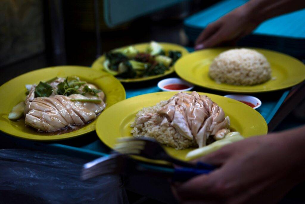 Yellow plates holding servings of Hainanese chicken rice, featuring succulent steamed chicken and rice, are arranged on a blue tray. A diner's hands are seen holding utensils near the meal, which is served with a side of green vegetables and small dishes of chili sauce.