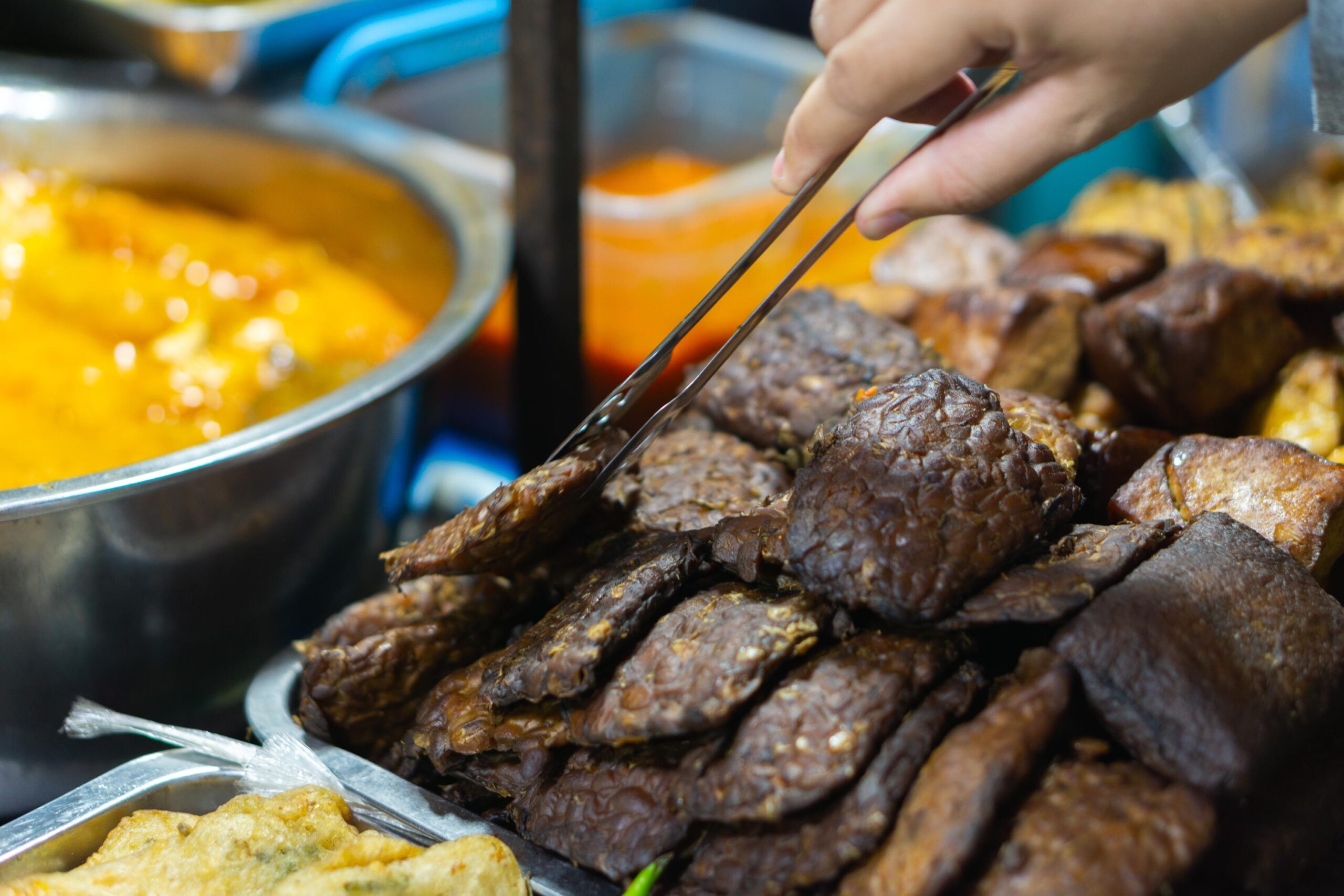 This close-up image captures a hand using metal tongs to select a piece of dark, fried tempeh from a generous stack. Adjacent to the savory bean cakes, a large metal bowl filled with a bright orange-yellow curry or sauce sits behind a tray of other golden-fried fritters.