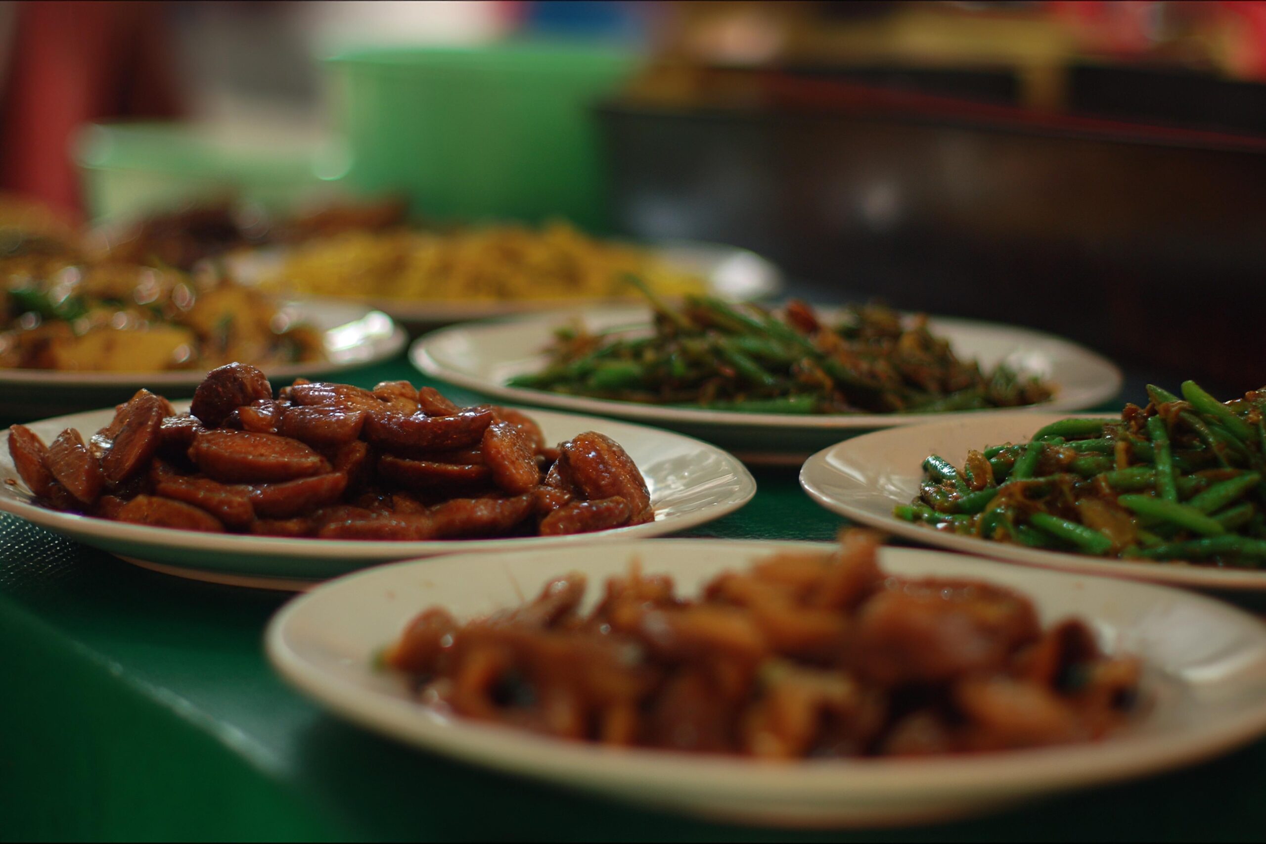 This close-up image presents a spread of freshly cooked dishes served on white plates, featuring glossy, reddish-brown sliced sausages and stir-fried green beans. The shallow depth of field draws attention to the texture of the meat in the mid-ground while softly blurring the surrounding plates of food on the green table surface.