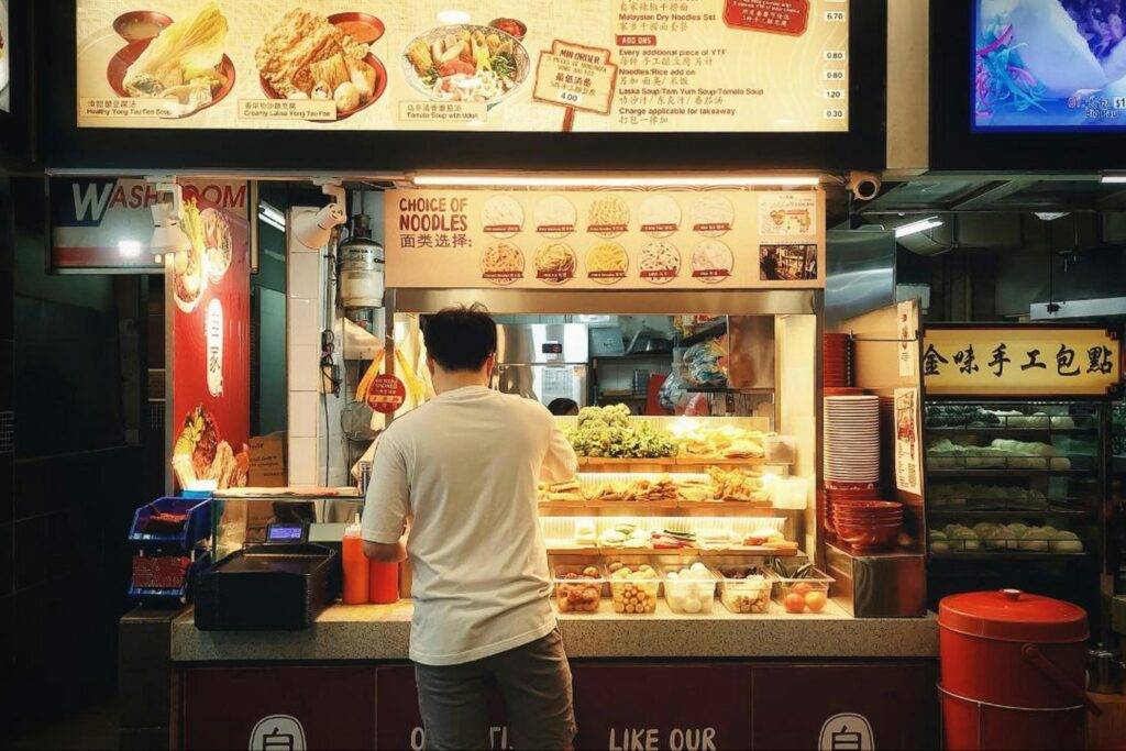A man in a white t-shirt stands at the counter of a brightly lit food stall, browsing through a variety of fresh ingredients and fried items displayed in trays. Above him, illuminated menu boards list noodle choices and prices, capturing the typical atmosphere of a bustling hawker center.