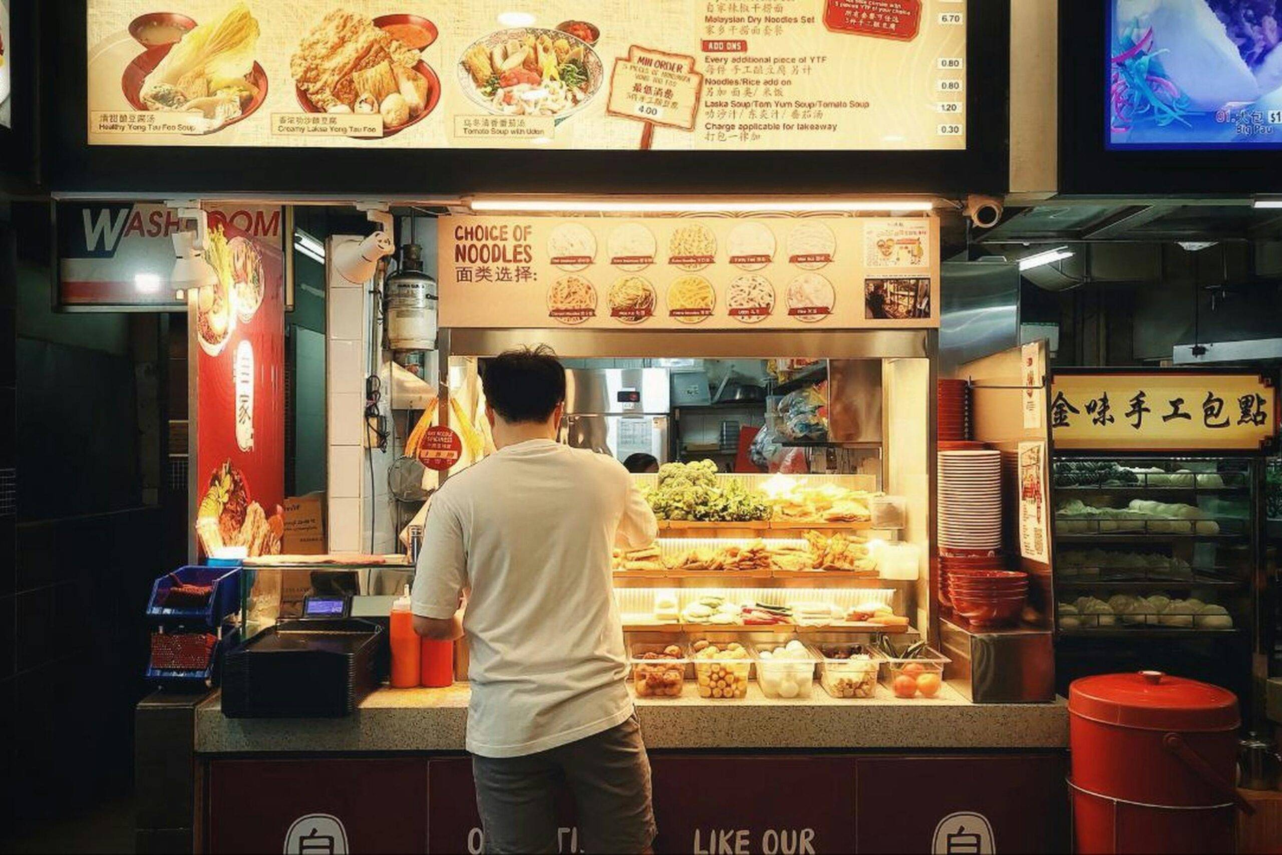 A man in a white t-shirt stands at the counter of a brightly lit food stall, browsing through a variety of fresh ingredients and fried items displayed in trays. Above him, illuminated menu boards list noodle choices and prices, capturing the typical atmosphere of a bustling hawker center.