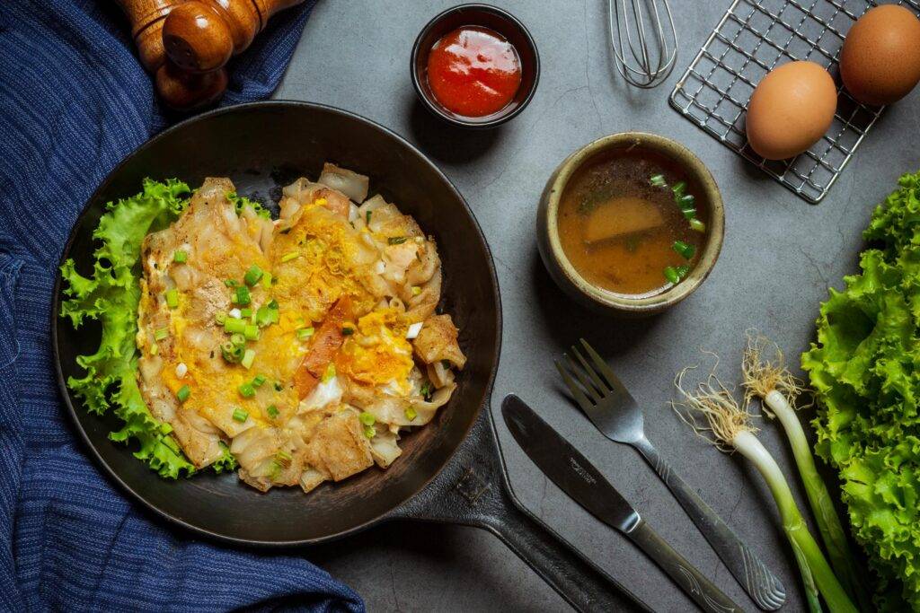A cast-iron skillet presents a serving of stir-fried wide rice noodles with egg and green onions, accompanied by a small bowl of soup and red dipping sauce. The overhead composition is framed by fresh ingredients, including lettuce, scallions, and raw eggs, along with silverware and a blue cloth set against a dark grey background.