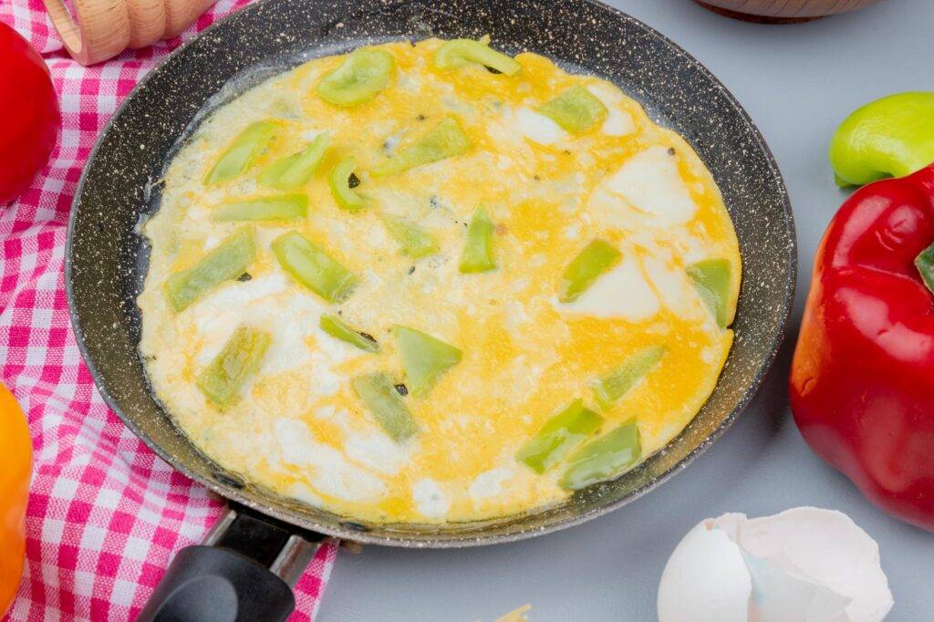 A speckled frying pan sits centrally in a top-down view, containing a cooked omelet topped with chunks of green bell pepper. The pan is surrounded by fresh red and yellow bell peppers, a red and white checkered cloth, and a broken eggshell on a plain grey surface.
