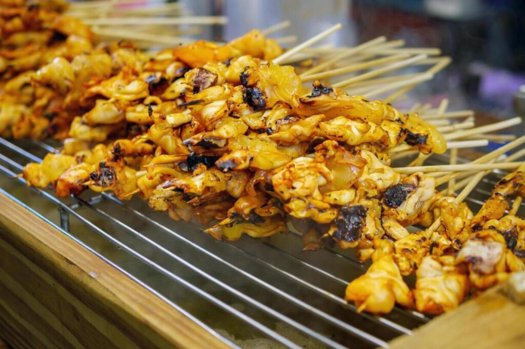 This close-up shot features a mouth-watering pile of golden-brown grilled skewers, likely chicken or satay, stacked high on a metal cooling rack. The meat is threaded onto bamboo sticks and shows appetizing char marks and a glossy glaze, hinting at the savory flavors found at this hawker center.