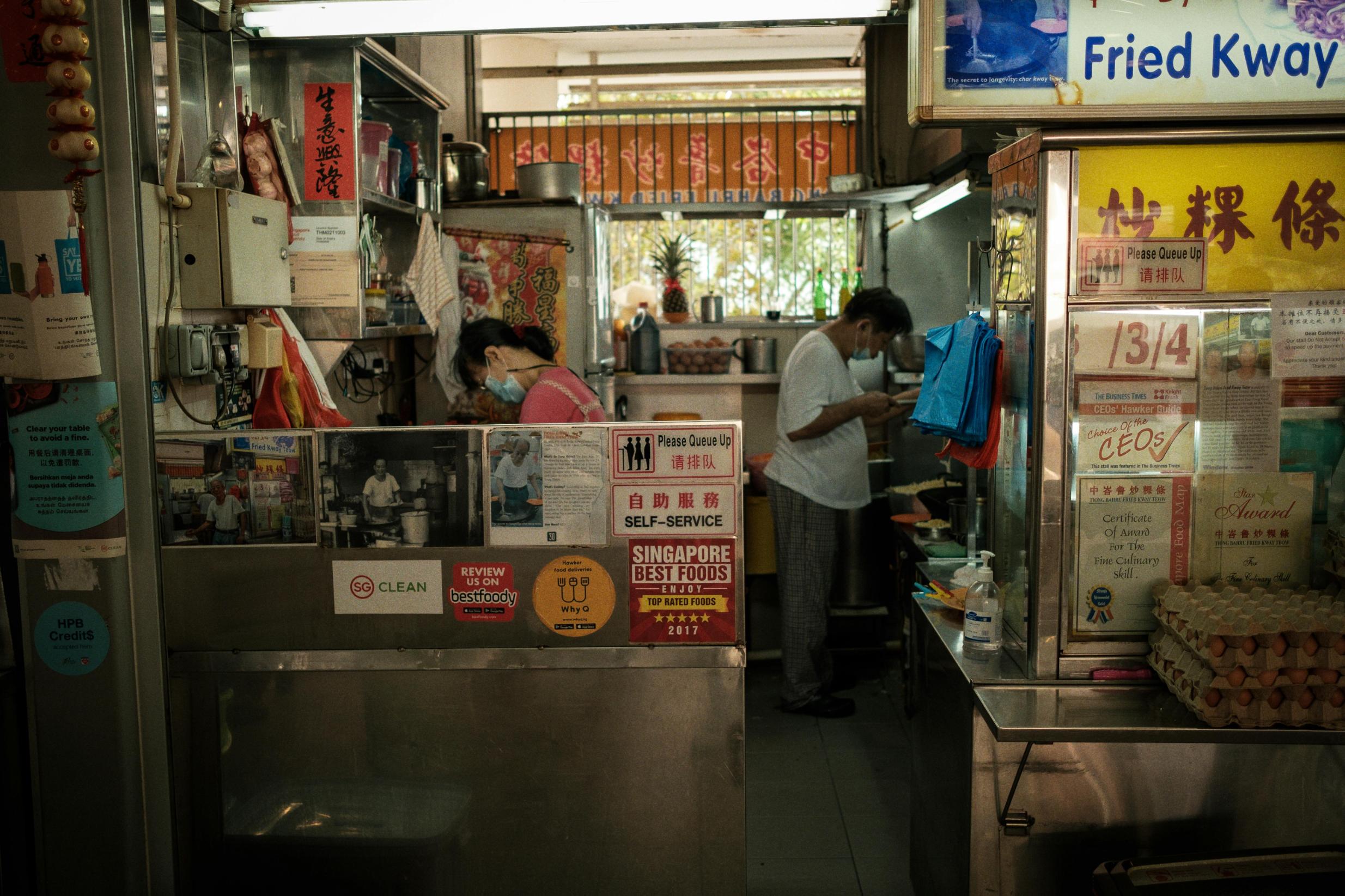 This candid shot captures the interior of a compact hawker stall where two vendors are busy at work amidst a clutter of cooking equipment and ingredients. The glass storefront is heavily decorated with accolades, newspaper clippings, and a "Fried Kway Teow" sign, sitting above stacks of egg trays ready for use.