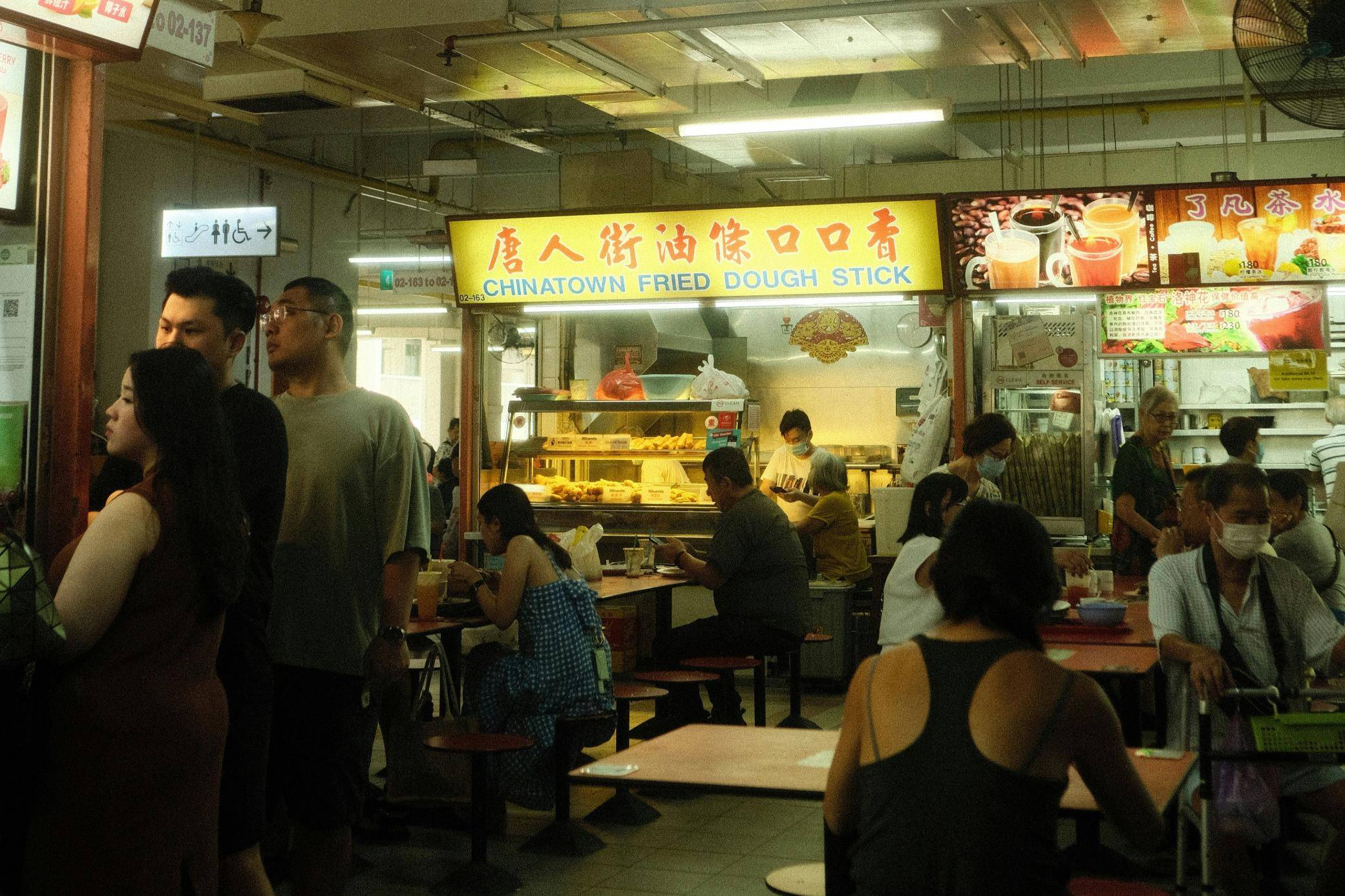 This image captures a lively scene at a bustling indoor hawker center or food court, featuring a central stall titled "Chinatown Fried Dough Stick." Numerous patrons are seen seated at tables or standing nearby, creating a vibrant atmosphere of everyday dining and community in a brightly lit urban setting.