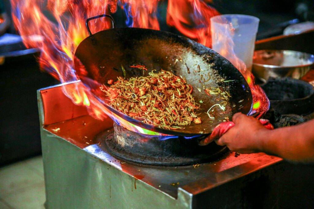 This image captures a dynamic action shot of a chef stir-frying noodles in a large wok over an intense, high-heat flame. The vibrant orange and blue fire engulfs the base of the pan, highlighting the "wok hei" technique used to infuse the dish with a smoky charred flavor.