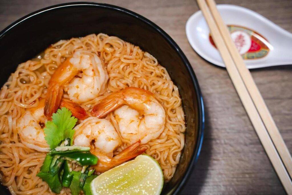 This image displays a bowl of noodle soup topped with large, succulent shrimp, fresh cilantro, and a wedge of lime for garnish. Beside the bowl sits a traditional ceramic spoon and a pair of wooden chopsticks, ready for a flavorful meal.