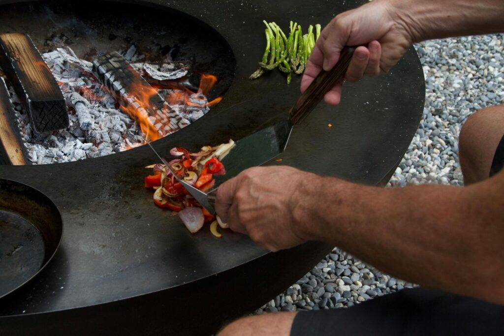 This image shows a person using two metal spatulas to grill sliced peppers and onions on the flat circular surface of an outdoor wood-burning fire pit. Nearby, a bunch of asparagus spears waits to be cooked over the embers, while the surrounding area is covered in light-colored pebbles.