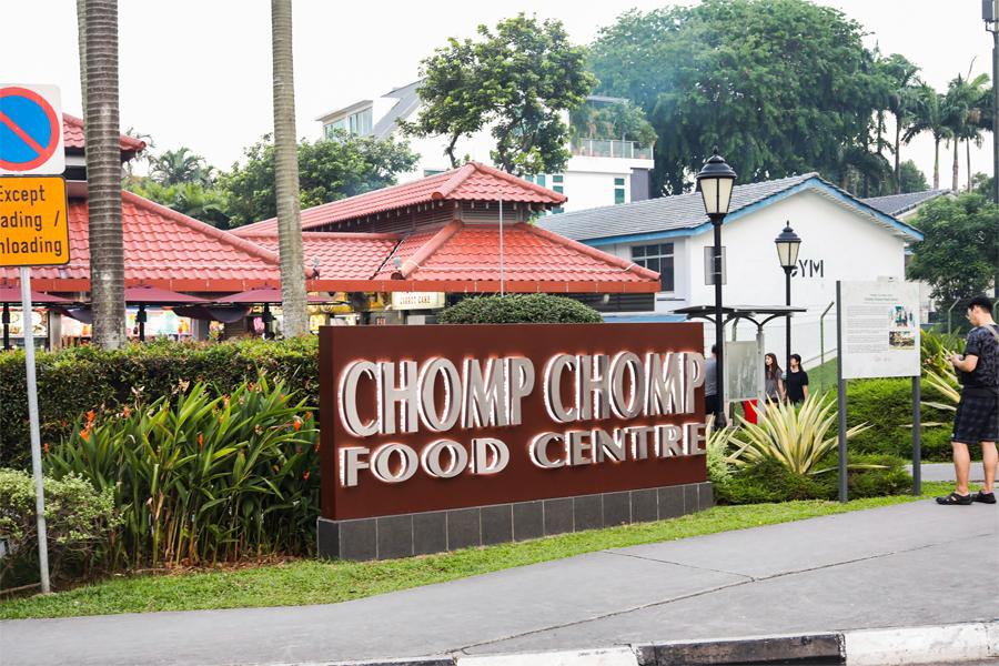 A large, rectangular sign prominently displaying "CHOMP CHOMP FOOD CENTRE" stands at the entrance, surrounded by lush green plants and trees. In the background, buildings with red-tiled roofs are visible, while pedestrians can be seen walking along the paved sidewalk to the right of the sign.