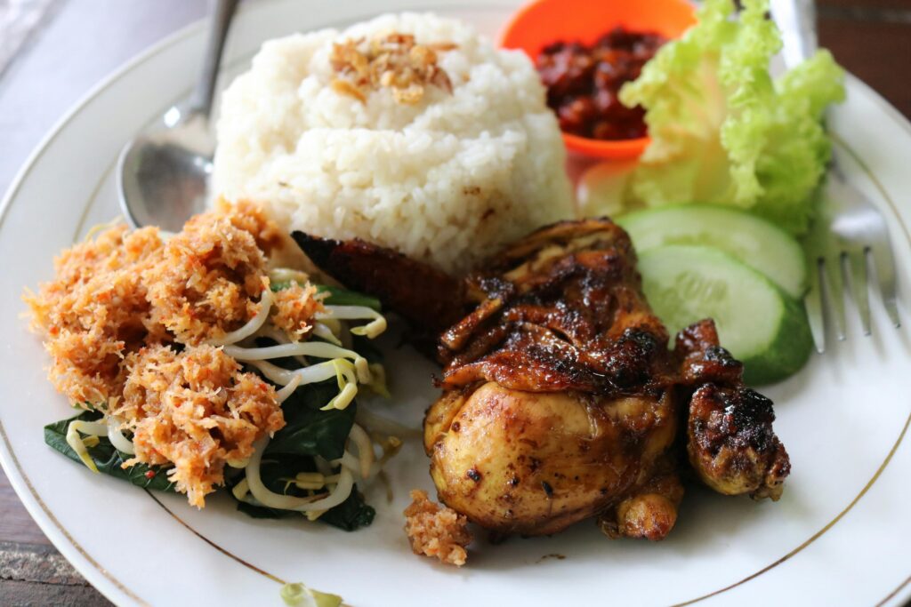 A white plate displays a meal consisting of white rice topped with fried shallots, a piece of roasted chicken, and vegetables garnished with a shredded orange topping. Fresh cucumber slices, lettuce, and a small side of red chili sambal round out the dish, which is served with a spoon and fork.
