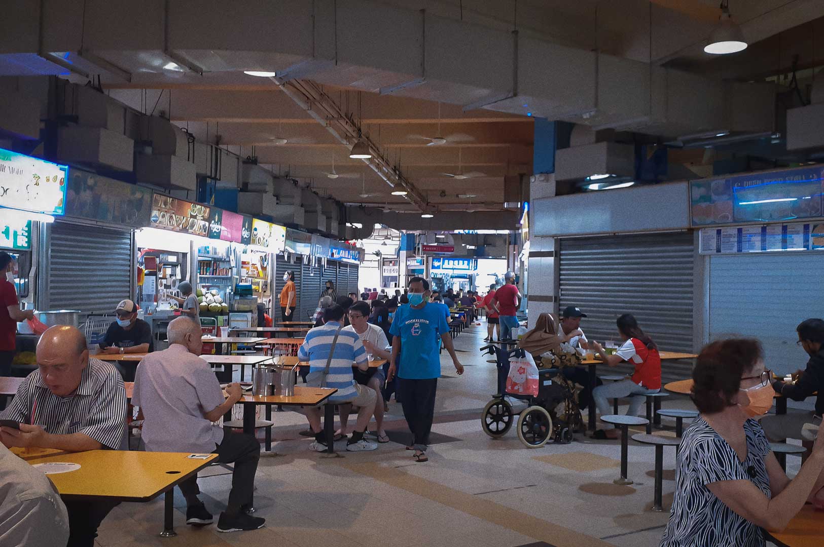 Indoor hawker centre with open food stalls, diners seated at yellow tables, and people walking through the central aisle under bright overhead lighting.