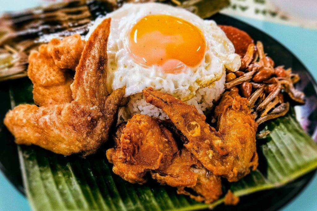 Close‑up of nasi lemak featuring crispy fried chicken wings, sunny‑side‑up egg, white rice, sambal, and fried anchovies served on a banana leaf.