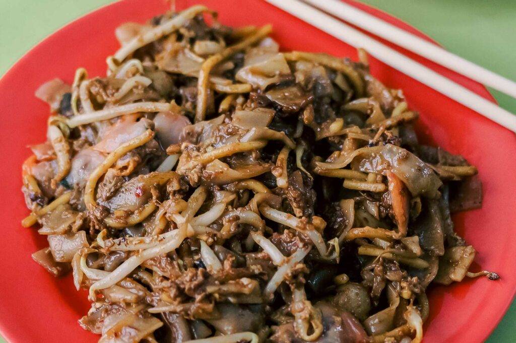 Close-up of kway teow mee on a red plate, featuring stir‑fried flat and yellow noodles with bean sprouts and savory toppings.