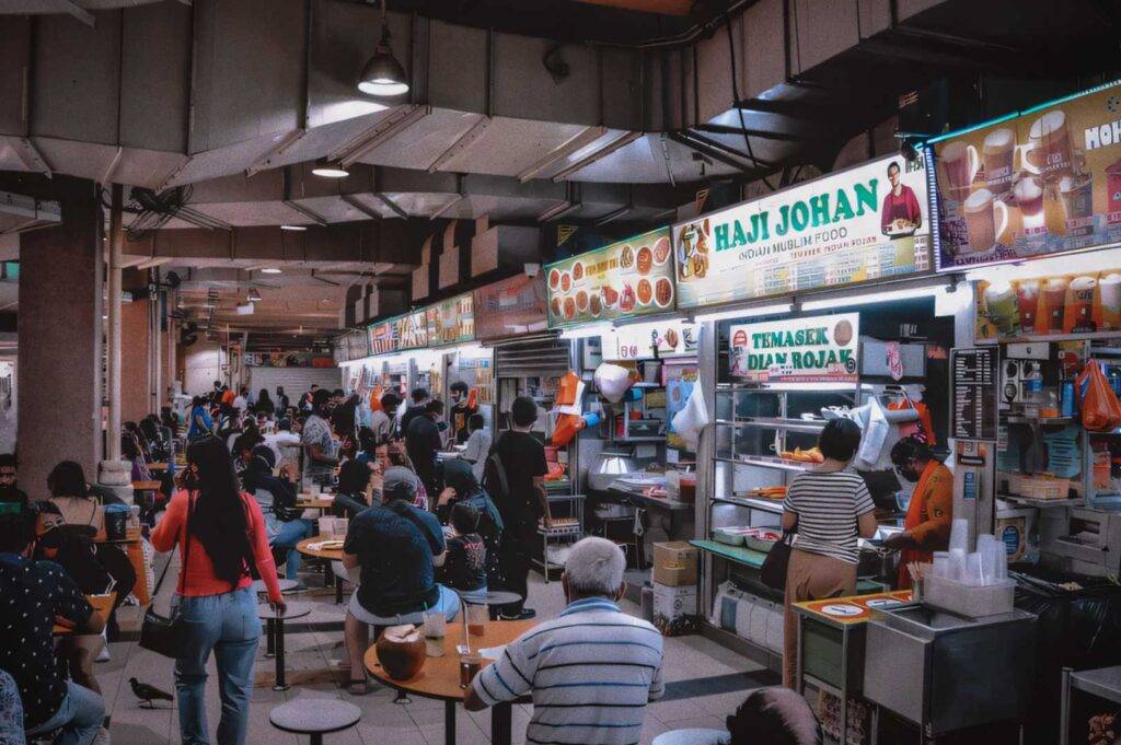 Busy hawker centre with diners seated at round tables and brightly lit food stalls serving Indian Muslim dishes and local snacks.