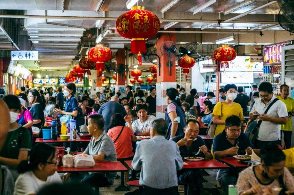 Crowded indoor hawker centre decorated with red lanterns, with many people seated at communal tables eating and others lining up at brightly lit food stalls.