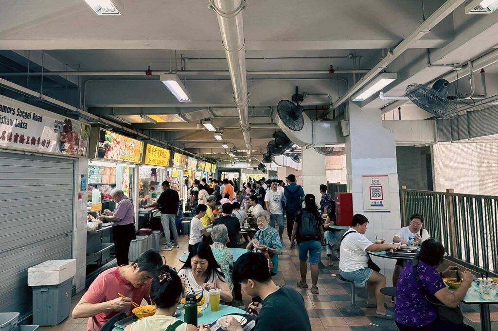 Crowded hawker centre with diners enjoying local Singapore food and vendors operating food stalls.