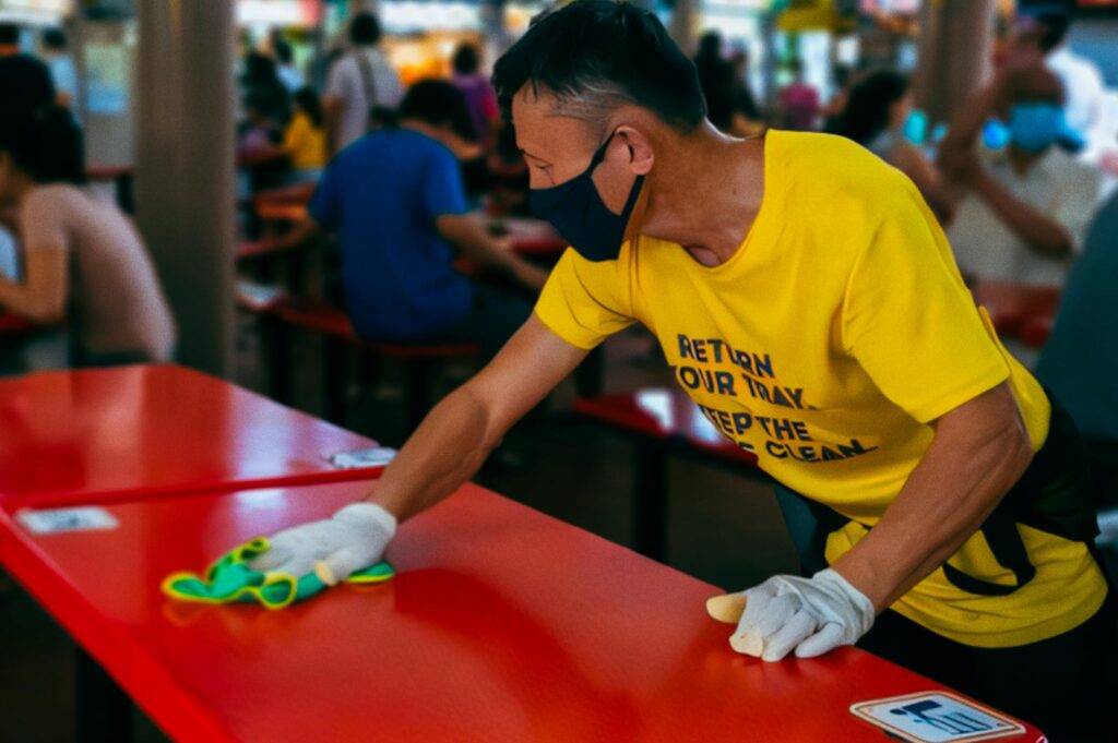 Staff member in a yellow shirt cleaning red tables at a busy hawker centre, wiping the surface with gloves and a cloth during routine upkeep.