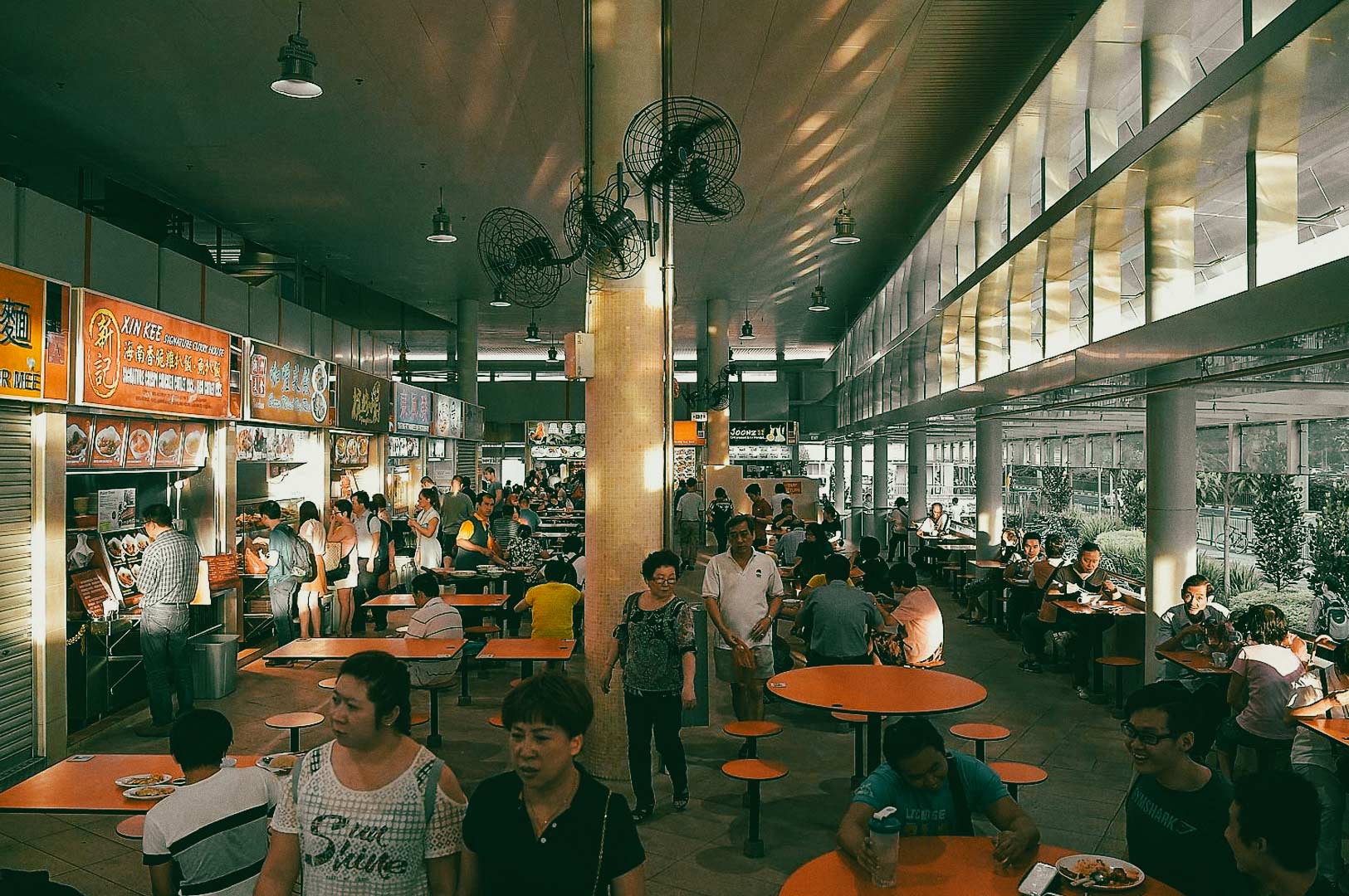 A busy hawker center with food stalls, orange tables, and people eating or walking through the open dining area.