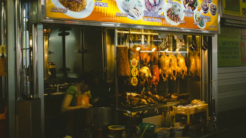 Close‑up shot of roasted meat hawker stall with ducks and pork hanging in glass display, vendor preparing food behind counter.