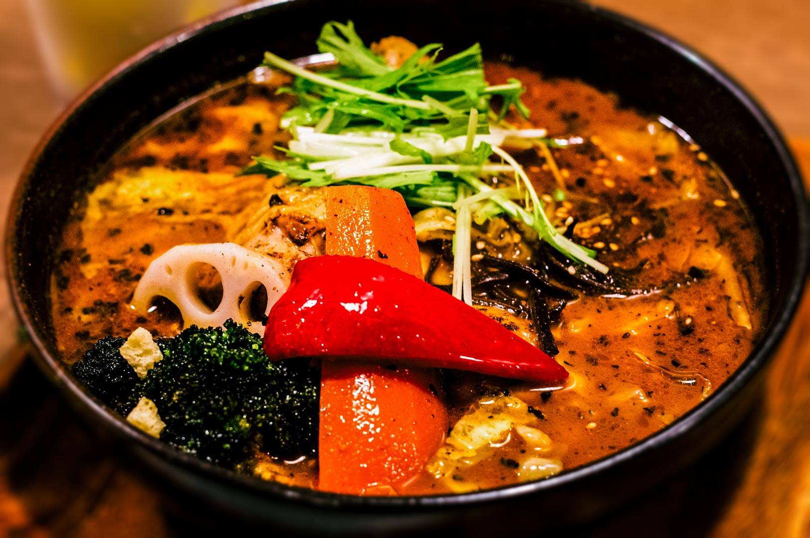 Bowl of Hokkaido soup curry filled with assorted vegetables including carrot, bell pepper, broccoli, lotus root, and greens in a rich, spiced broth.