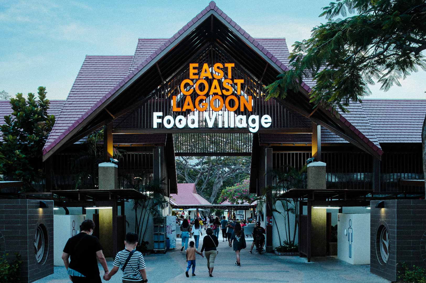 Evening view of the East Coast Lagoon Food Village entrance, featuring the large illuminated sign and people walking into the open-air dining area surrounded by trees.