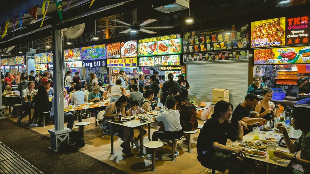Wide-angle street-level shot of a bustling Asian hawker centre at night, showing rows of food stalls with illuminated menus and diners seated at shared tables enjoying local street food.