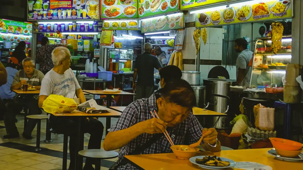 Eye‑level shot of bustling hawker center with man eating at table, Indian food stall menus glowing in background.