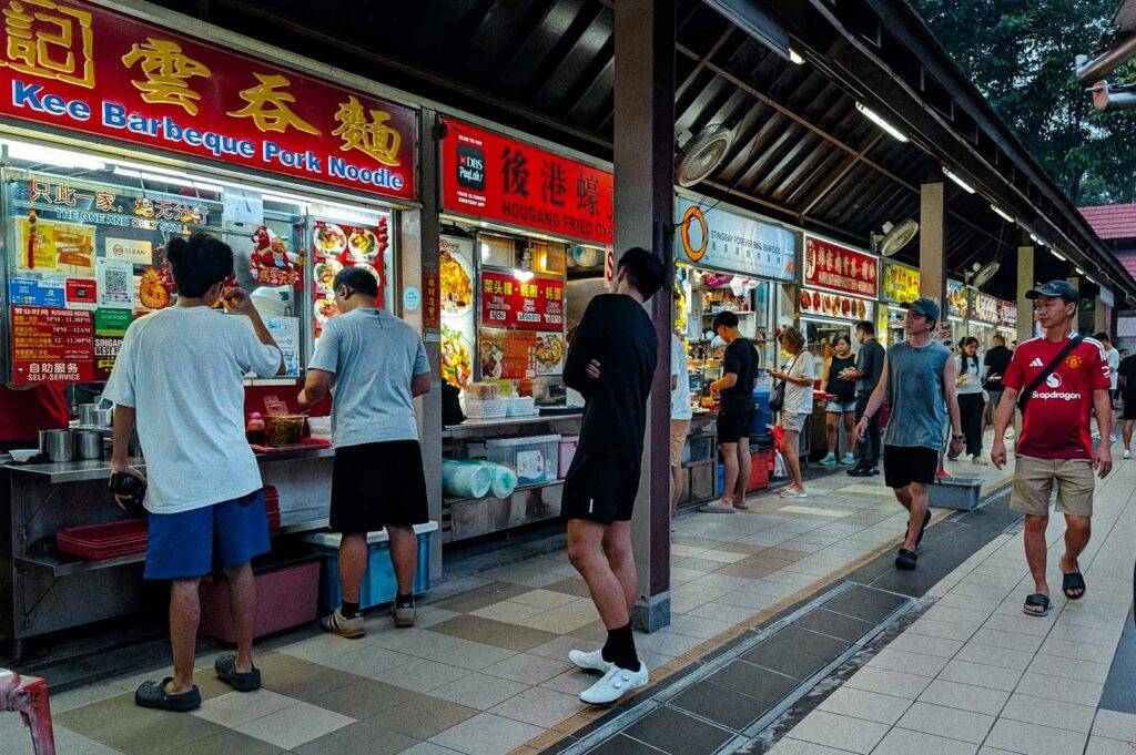 Row of brightly lit hawker stalls at East Coast Lagoon Food Village, including Kee Barbeque Pork Noodle and other Asian food vendors, with customers ordering at the counters.