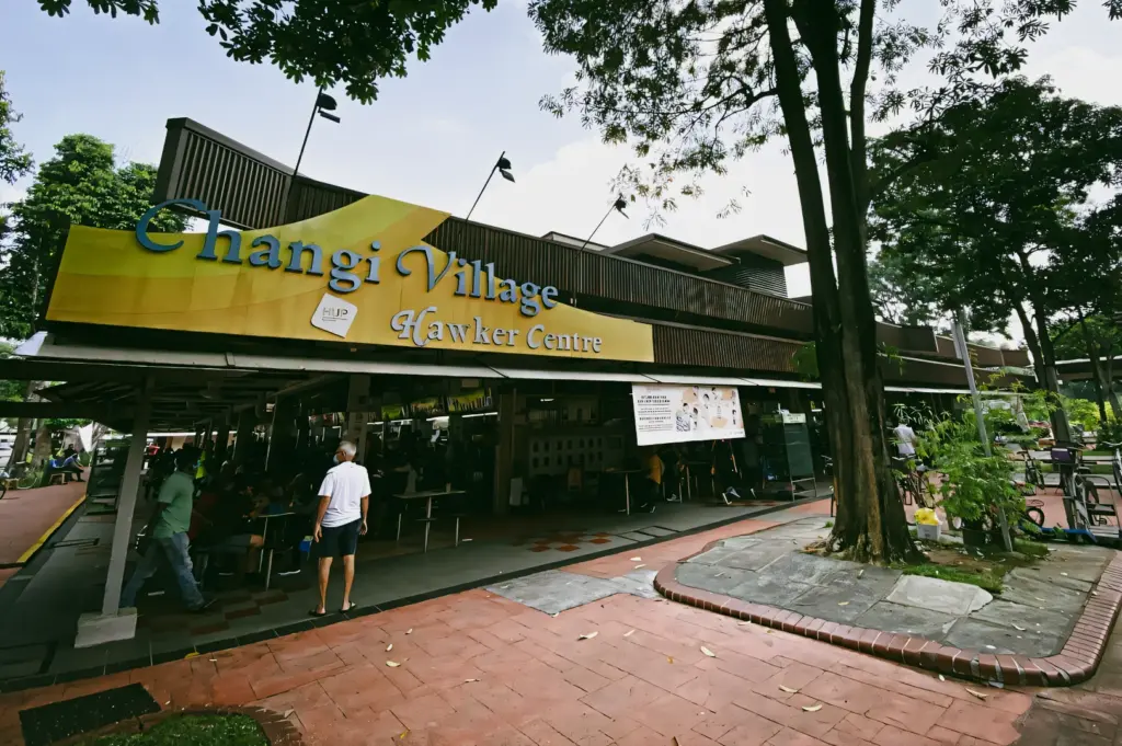 Exterior of Changi Village Hawker Centre with its yellow signage, surrounded by trees and outdoor seating along a paved walkway.