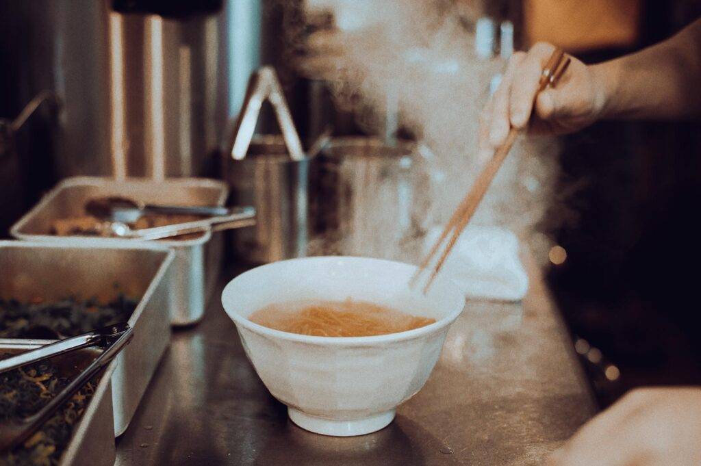 Bowl of hot noodles being prepared in a kitchen with steam rising.