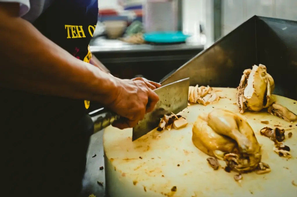 Close-up eye-level shot of a cook slicing poached chicken on a chopping board with a cleaver, showing freshly cut pieces and food preparation in a commercial kitchen environment.