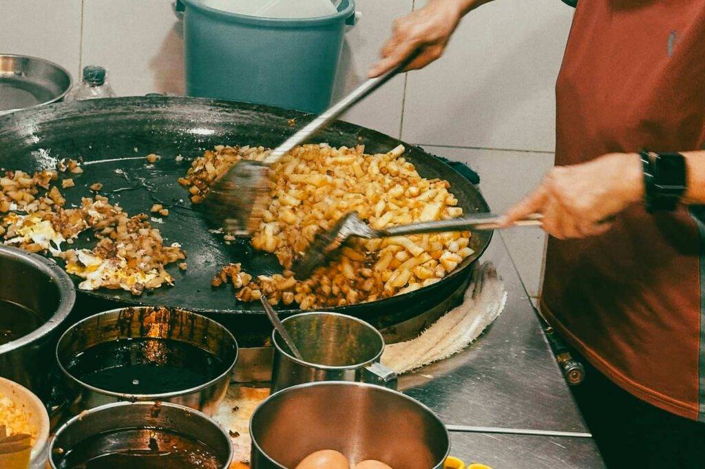 Hands cooking diced carrot cakes in a large flat wok, using metal spatulas to stir the ingredients, with bowls of sauces and eggs arranged around the cooking station.