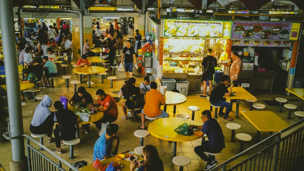 Wide‑angle shot of crowded Southeast Asian hawker center with diners at yellow tables, food stalls offering Indonesian cuisine in background.
