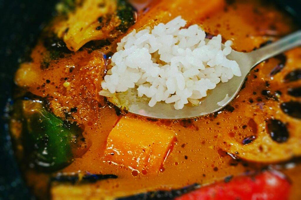 Close‑up of a spoonful of white rice held above a bowl of Hokkaido soup curry with vegetables in a flavorful, reddish broth.