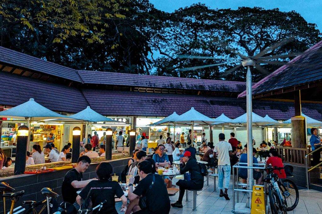 Busy outdoor dining section at East Coast Lagoon Food Village during the evening, with diners seated under large fans and umbrellas near various hawker stalls.
