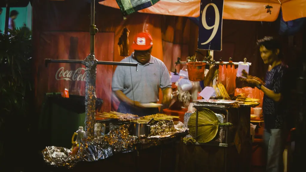 Close‑up angle of skewers grilling on foil‑covered charcoal, two cooks preparing food at bustling night market stall.