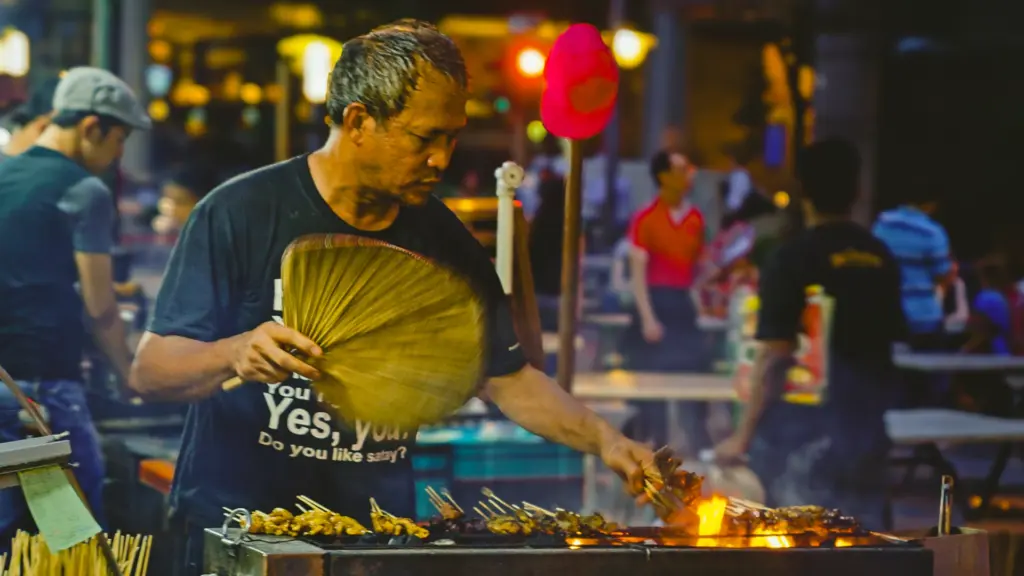 Medium‑angle shot of street food vendor fanning flames over satay skewers at night market, wearing playful satay T‑shirt.