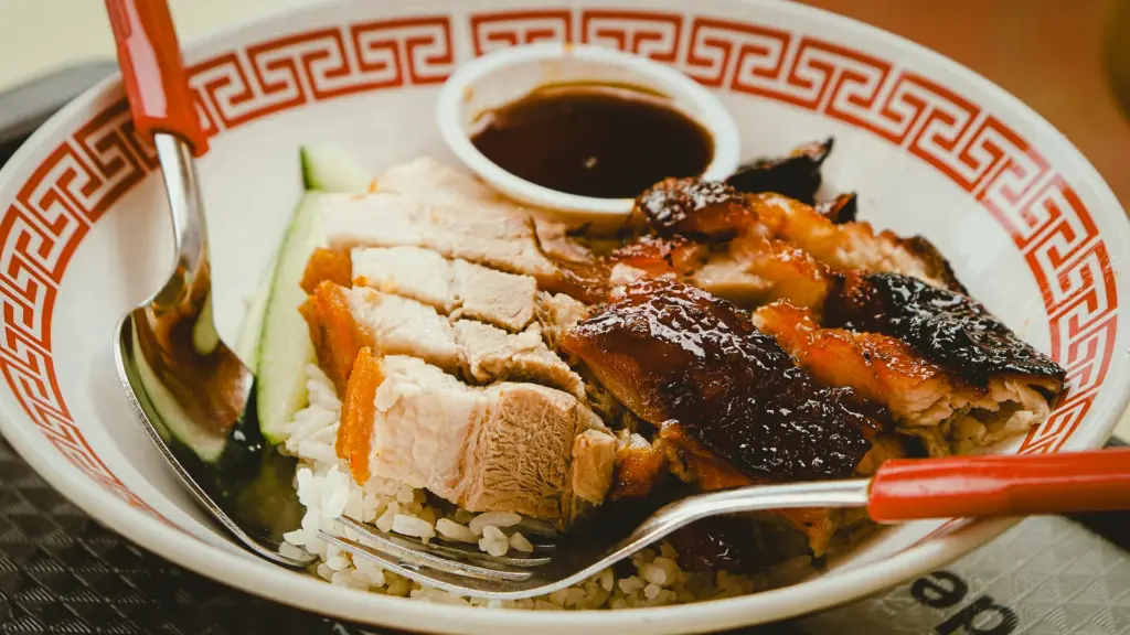 Close‑up angle of Asian rice bowl topped with roasted pork and chicken slices, cucumber garnish, and dipping sauce.